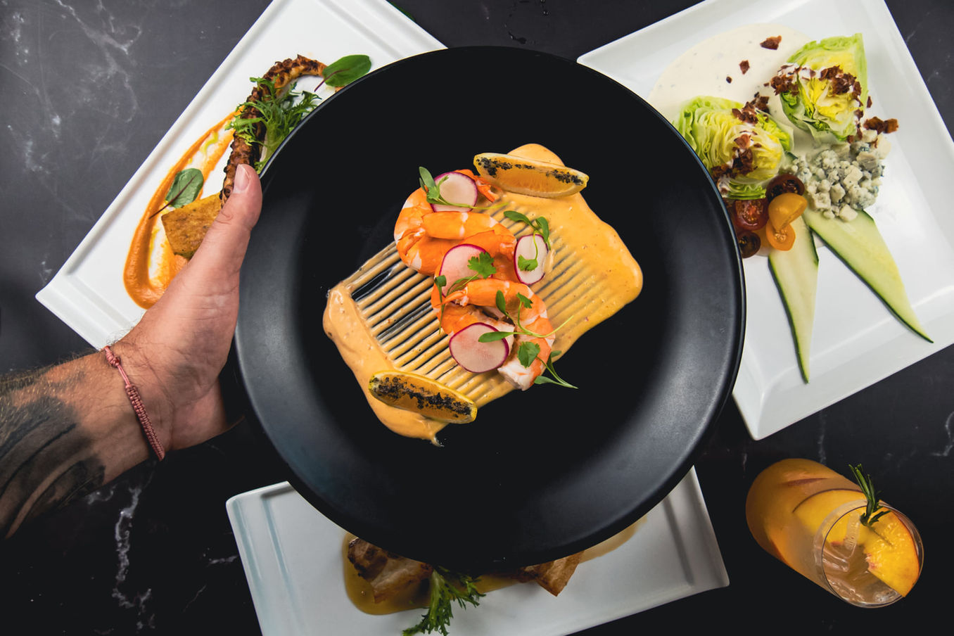 a man's hand holding a shrimp dish on a black plate up over other dishes and a cocktail on a table at Milagro Restaurant & Bar
