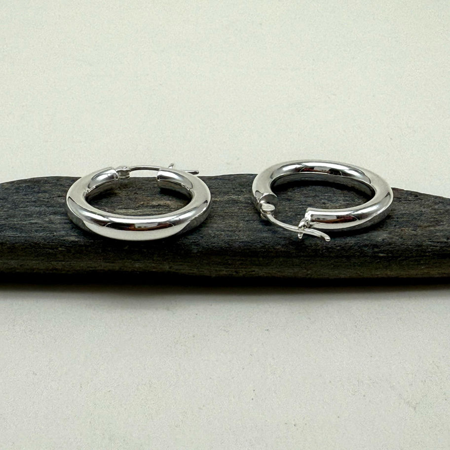 Polished silver Tube Hoop Earrings displayed side by side on a dark stone surface.