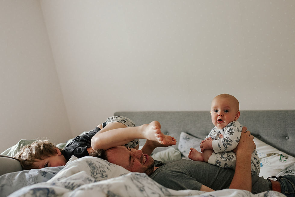 en pappa ligger i sängen och håller i ett spädbarn medan ett äldre barn klättrar på hans huvud och gör honom illa