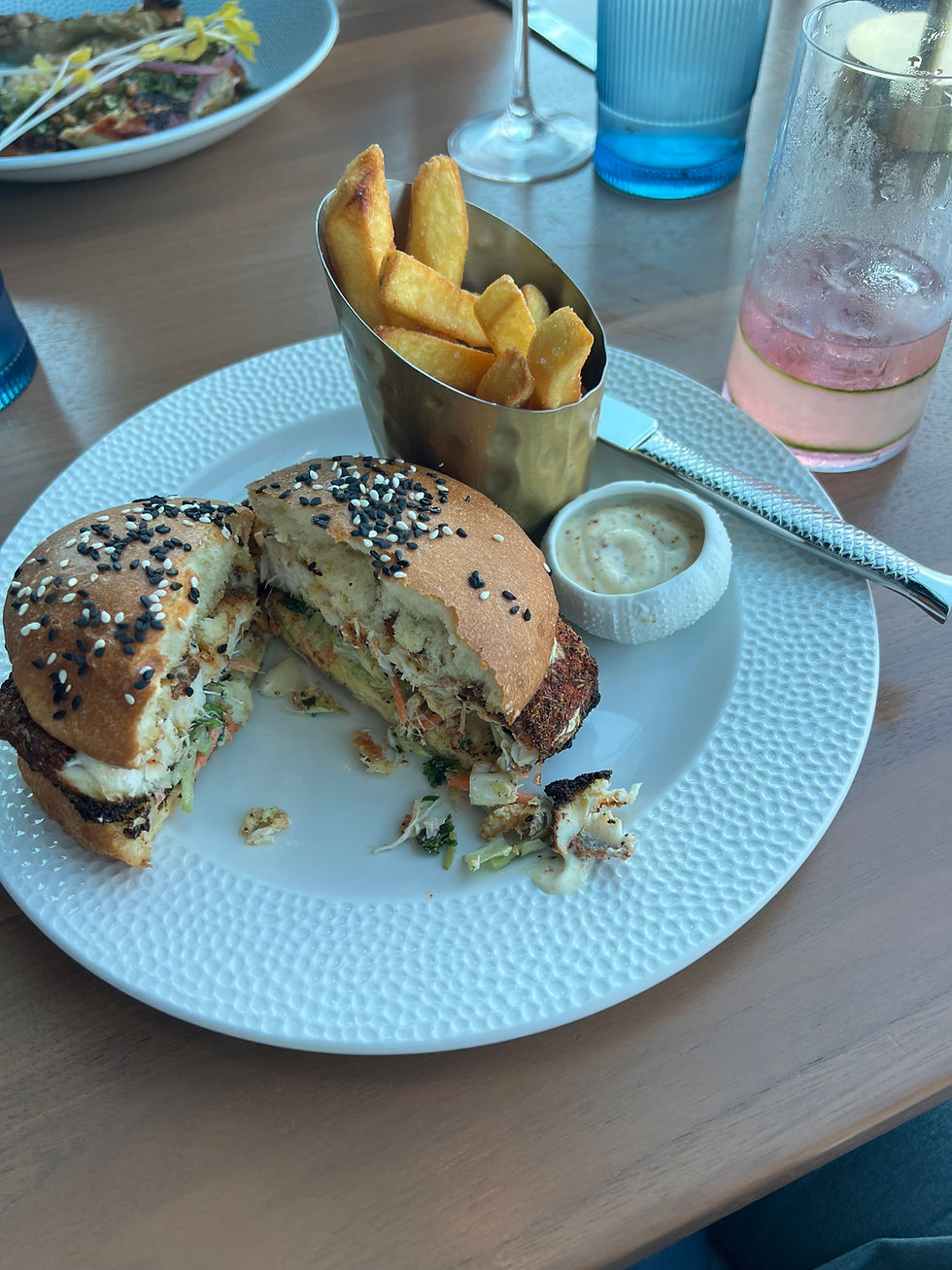 Burger with sesame bun, fries in a metal cup, and a drink on a wooden table. Plate has white textured design. Relaxed dining scene.