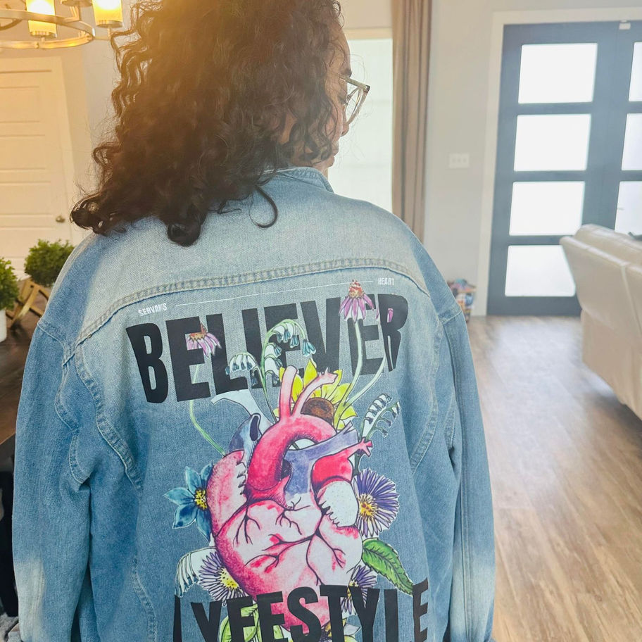 Woman wearing denim jacket with the word BELIEVER and heart graphic.