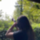 A person with long hair aims a shotgun toward the sky in a lush, green outdoor setting surrounded by trees and foliage.