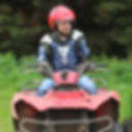 A man wearing a red helmet and blue protective gear rides a muddy red ATV outdoors, with green foliage in the background