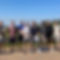 A group of ten men stand outdoors in a line on a gravel path, some smiling, some wearing sunglasses. Two men have medals.