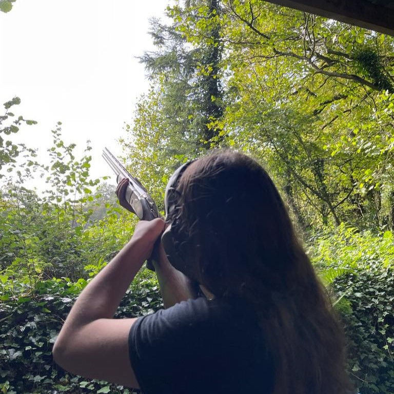 A person with long hair aims a shotgun toward the sky in a lush, green outdoor setting surrounded by trees and foliage.