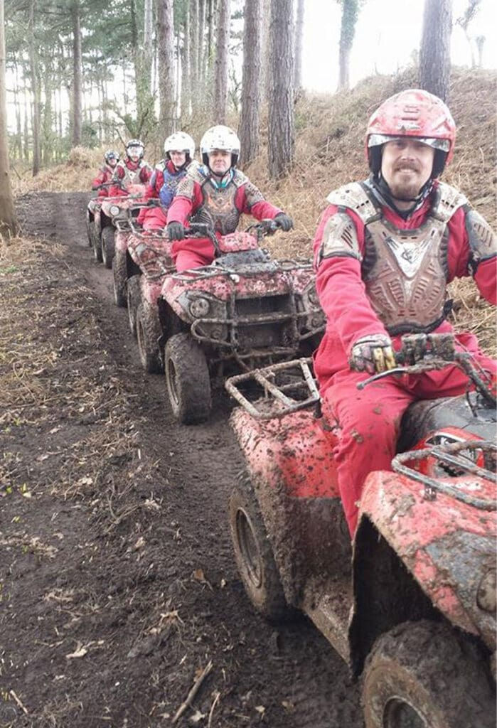 Five people in red protective gear and helmets ride muddy quad bikes in a single file on a dirt path through a wooded area