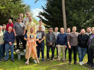 Group of smiling men posing outdoors in a wooded park, some holding medals, with an inflatable doll in front