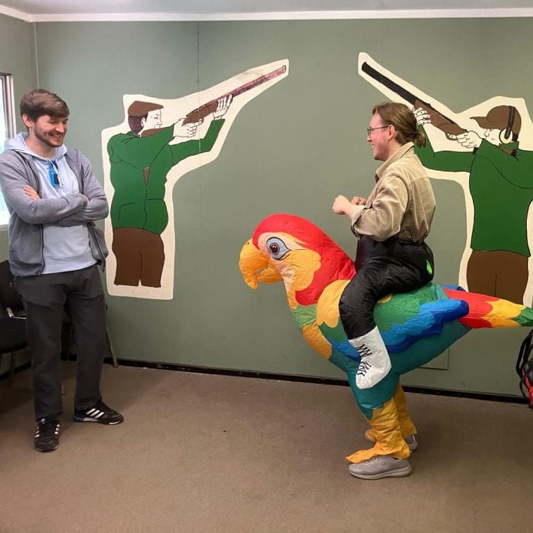 Man rides a colorful parrot costume while a smiling man watches; green wall behind has shooter cutouts.