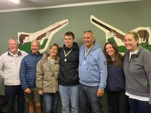 Smiling group of seven wearing medals poses indoors before a green wall with shooting silhouettes.