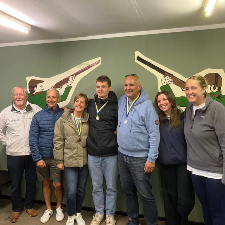Smiling group of seven wearing medals poses indoors before a green wall with shooting silhouettes.