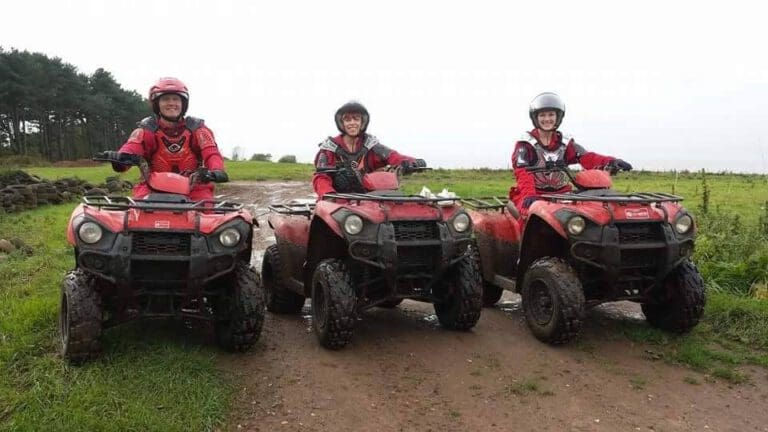Three people wearing helmets and red jackets sit on muddy red quad bikes, smiling and posing on a dirt path