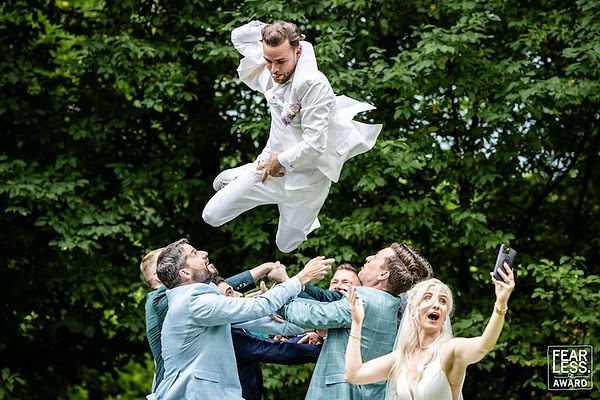 Bräutigam wird von seinen Freunden mit vollem Einsatz in die Luft geworfen, während die Braut lachend ein Selfie macht – ein preisgekrönter, unvergesslicher Hochzeitsmoment voller Freude und Action im Grünen. Fearless Award Siegerfoto von Mischa Baettig.