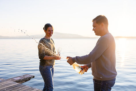 Ein Mann öffnet Champagner, während eine Frau zwei Gläser am Seeufer bei Sonnenuntergang f