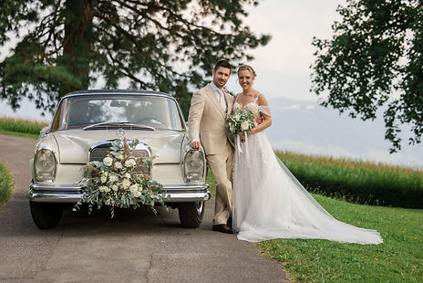 Das Brautpaar steht bei einem Oldtimer mit Blumen vor Schlössli Utenberg Luzern, umgeben v
