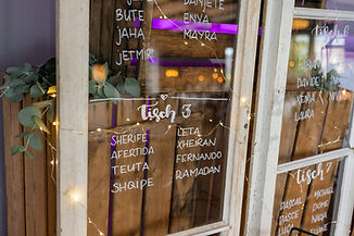 Rustikaler Sitzplan auf Fensterrahmen mit weißen Namen, Holz, Lichterkette und Grün. Besch