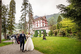 Das Brautpaar schreitet Hand in Hand vor dem Vitznauerhof am Vierwaldstättersee, gefolgt v