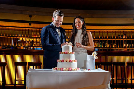 Ein frisch verheiratetes Paar schneidet gemeinsam eine dreistöckige Hochzeitstorte an der Bar im Theater Casino Zug an. Die Torte ist mit rosa und cremefarbenen Macarons verziert, und der Hintergrund zeigt eine beleuchtete Bar mit Flaschenregalen.