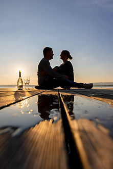 Ein Paar sitzt eng umarmt auf einem Holzsteg am See bei tief stehender Sonne. Daneben stehen eine Champagnerflasche und zwei Gläser. Eine Pfütze im Vordergrund spiegelt das warme Licht für eine romantische Stimmung