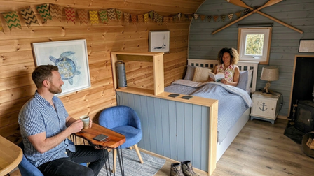 Interior photo of the Ayr glamping pod at Emlyn's Coppice. A man sits at a live edge sycamore table with a drink, whilst a woman lies in bed reading a book.