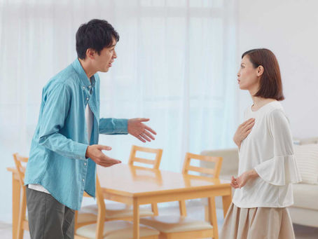 Man gestures while arguing with woman in a bright living room; she clutches her chest, looking upset.