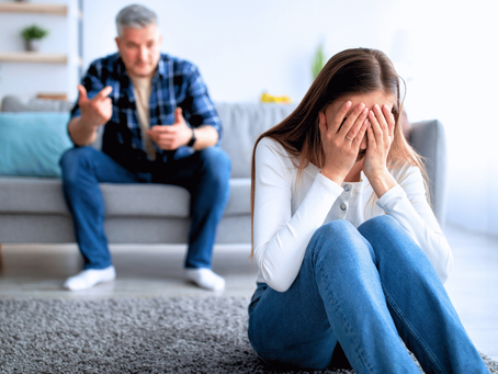 Distressed woman covers her face on the floor while a man argues on a sofa in a bright living room.