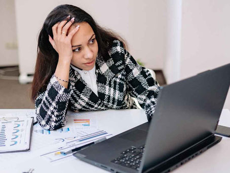 Woman in a checkered jacket sits at a desk looking stressed at a laptop. Charts and papers are scattered around in a bright office.