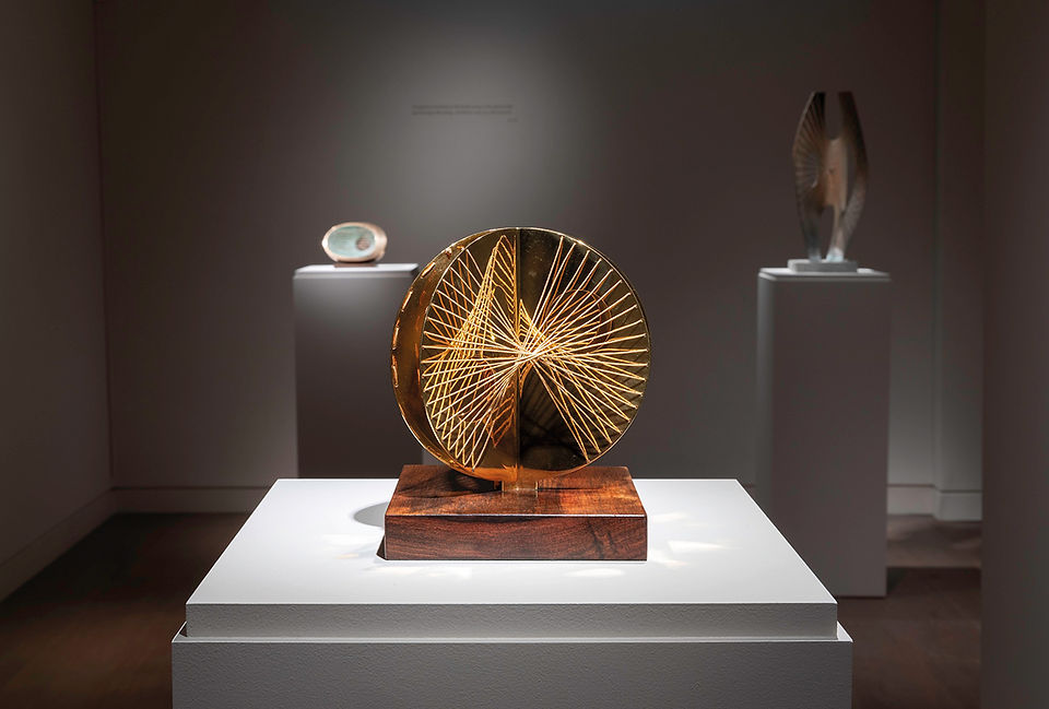 Abstract gold wire sculpture on a wooden base in a dim gallery, with two similar artworks on pedestals behind it. Installation photographs of Barbara Hepworth: Strings at Piano Nobile 