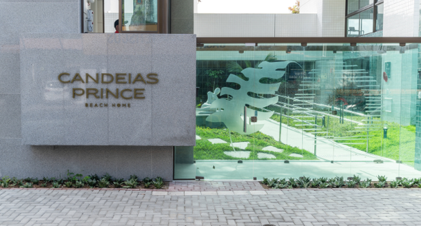 Acesso frontal ao edifício Candeias Prince com calçada acessível, projeto de paisagismo moderno, guarita de segurança e detalhes em vidro.