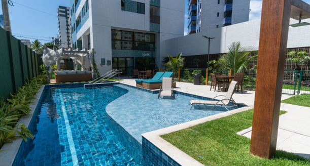Outro ângulo da área da piscina, destacando o deck de madeira premium, o paisagismo tropical ao redor e a integração com as áreas comuns.