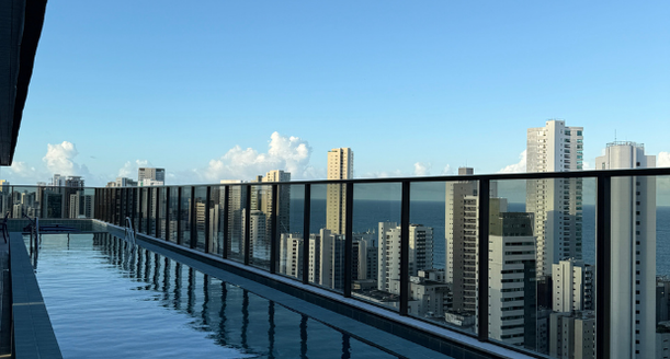 Piscina de alto padrão fotografada durante o entardecer, com iluminação interna acesa refletida na água e vista panorâmica da cidade ao fundo.