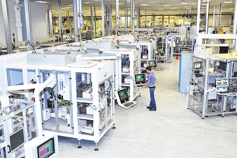 Worker monitors automated machinery in a bright factory, surrounded by white production lines and glowing control screens.