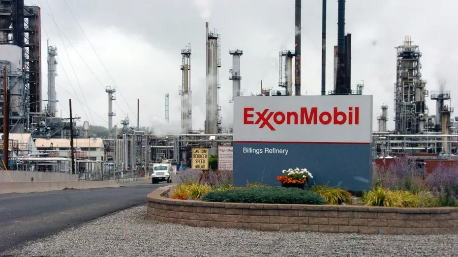 ExxonMobil Billings Refinery sign in front of smoky industrial towers and pipes at a large refinery complex.