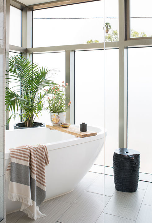 A serene and stylish master bathroom in a contemporary loft located in Atwater Village. This tranquil retreat features a sleek, white freestanding bathtub positioned next to floor-to-ceiling windows that frame picturesque views of palm trees. The natural light floods the space, highlighting the minimalist décor that includes a live potted palm and a delicate floral arrangement. Practical yet chic accessories, such as a wooden bath tray and a soft striped towel, add functionality and a touch of warmth. A textured black stool serves as a versatile accent piece, further enhancing the room’s modern, airy vibe.