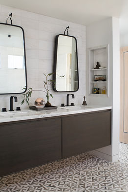 Stylish primary bathroom in a Playa del Rey coastal home featuring a double vanity with dark wood cabinets, marble countertop, unique black framed mirrors, and geometric patterned floor tiles, complemented by built-in shelving with elegant decor.