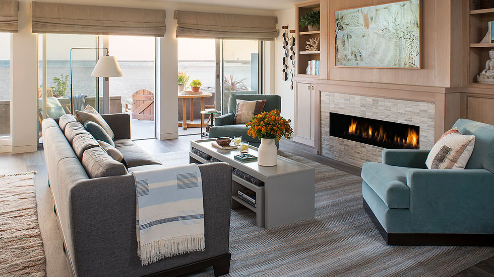 Cozy living room with gray sofas, a blue chair, a fireplace, and ocean view. A table with flowers centers the room, creating a warm ambiance. A beach house in Los Angeles.