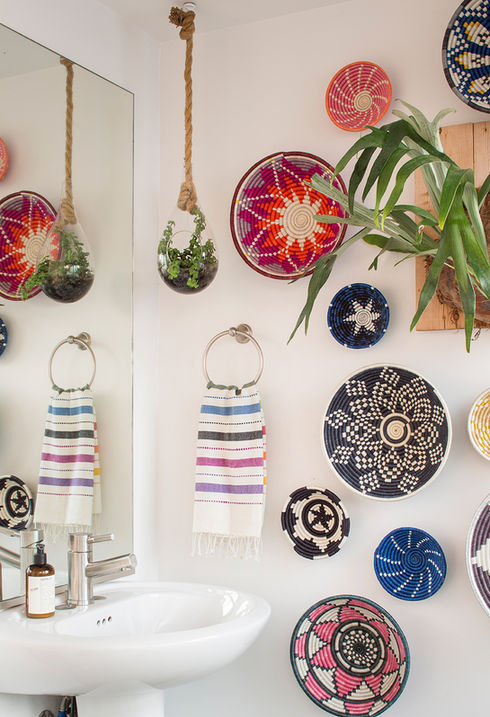 A bohemian-style bathroom featuring a white pedestal sink. The walls are adorned with colorful woven baskets in various patterns and sizes, complemented by hanging green plants in clear glass orbs with rope. A wooden shelf adds a natural touch. This decor gives the room a vibrant, contemporary, and art-filled look, typical of a loft in Atwater Village.