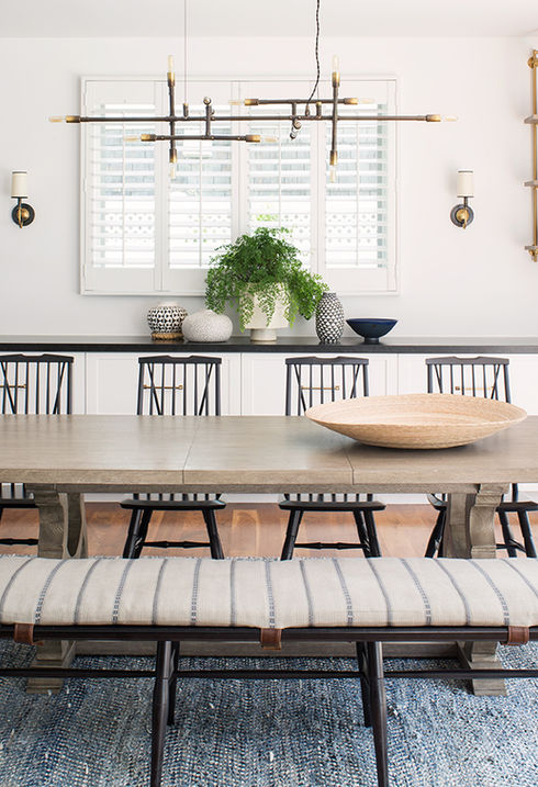 Charming dining room in a Pacific Palisades coastal home, featuring an oak dining table with matching bench and chairs, set on a textured blue rug, complemented by stylish wall shelves displaying decorative items and a modern chandelier above.