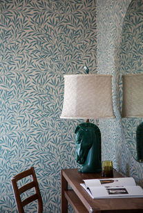 Home office with blue botanical wallpaper, green ceramic horse lamp, wood desk, and curved wall mirror.