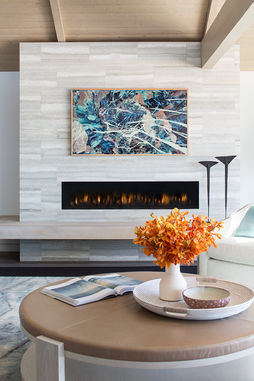 Cozy corner of a coastal living room in Playa del Rey, Los Angeles, highlighting a linear fireplace set in travertine tile with a vibrant abstract painting above and elegant fall-themed decor on the coffee table.