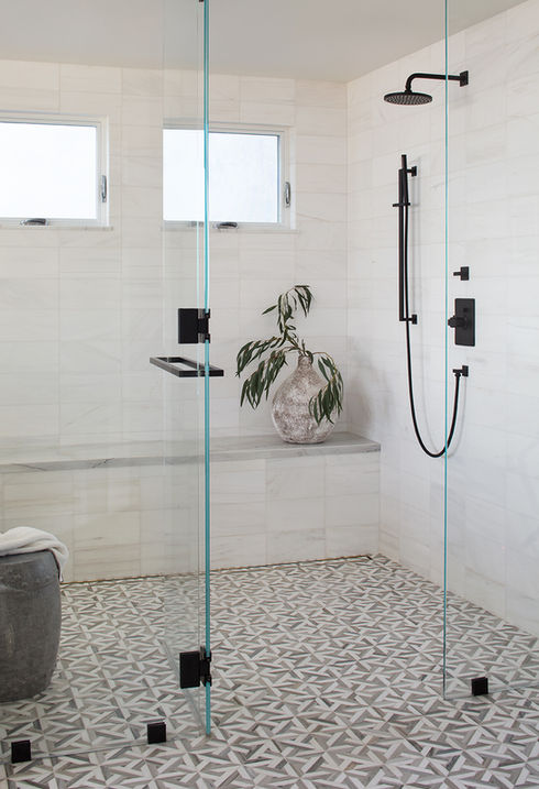 Elegant walk-in shower in a Playa del Rey coastal home's primary bathroom, featuring white marble tiles, a striking geometric floor, sleek black shower fixtures, and a glass enclosure, enhanced with a decorative plant for a touch of nature.