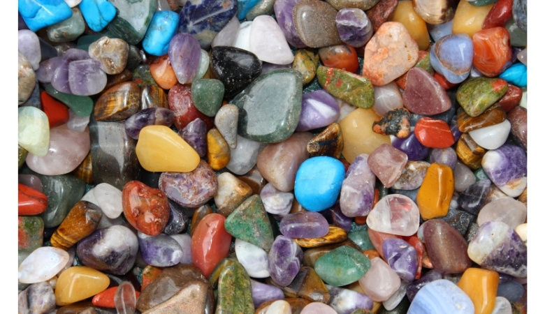 Colorful polished gemstones in various sizes form a vibrant mosaic. The stones are in blues, purples, reds, greens, and yellows.