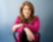 A woman dressed in professional attire is seated on a chair backwards, with her arms resting on the back.