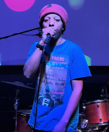 Kiidflexx, a member of Fallen7, performing on stage with a microphone, wearing a pink beanie and a blue shirt, with a drum set in the background.