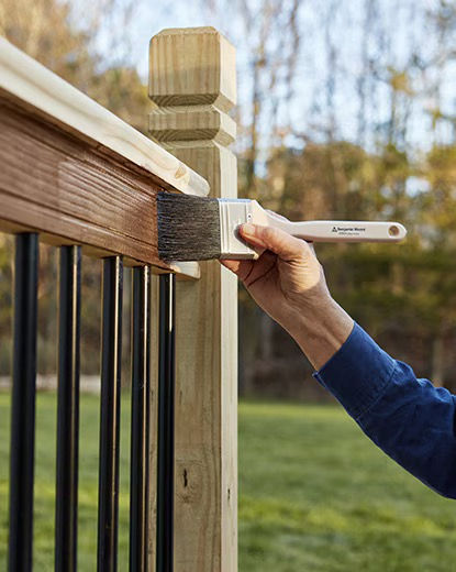 Hand in a blue sleeve stains a wooden deck railing with a brush outdoors, with yard and trees blurred behind.