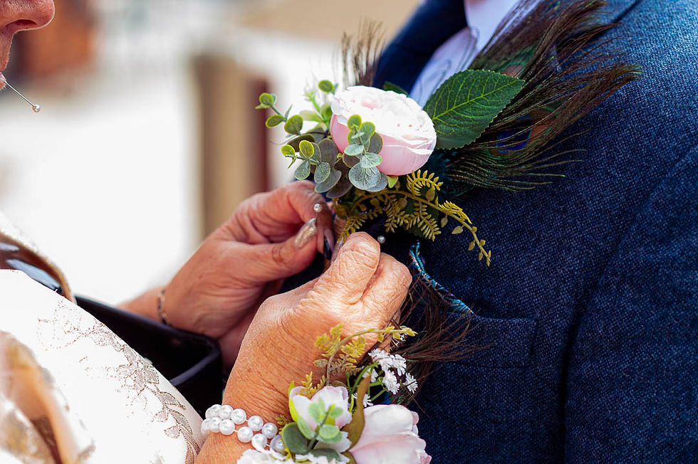 Hands pinning a pink rose boutonniere on a blue suit. The setting is bright, with a pearl bracelet and floral corsage visible. Elegant mood.
