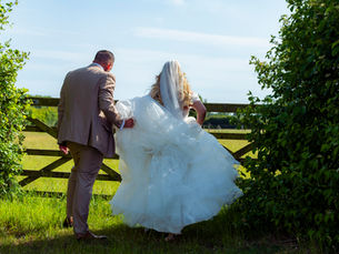 A bride in a white gown and veil, and a groom in a beige suit walk through a field gate surrounded by greenery under a clear blue sky.
