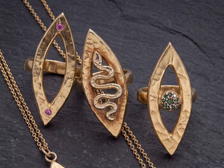 Three golden Vesica Piscis shaped rings. One with a serpent moving up the middle, one with a pink sapphire set at each point, one with a blue diamond iris and a black diamond pupil.