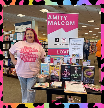 Indie author Amity Malcom stands in front of a display of her works wearing a pink shirt.