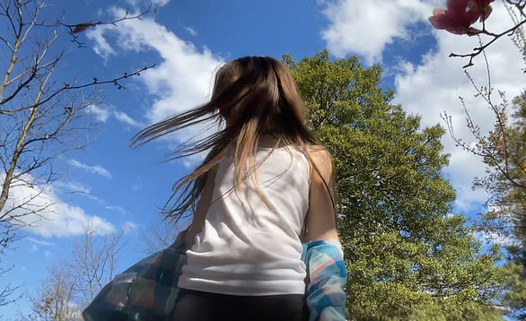A girl with long brown hair faces away under a blue sky with scattered clouds. Trees and blooming branches are behind her.