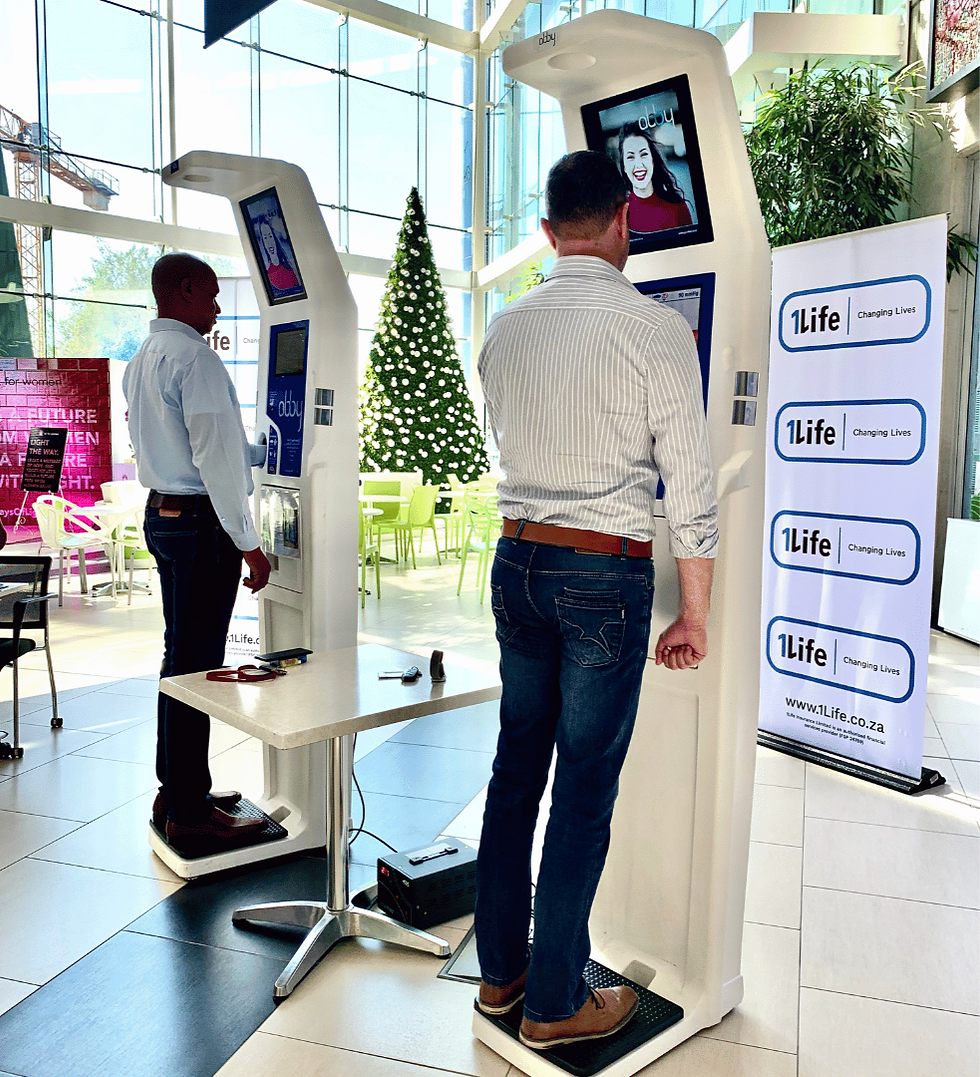 Two men use health kiosks indoors in a workplace, near a decorated Christmas tree. A smiling woman's photo is on the screen. Banner reads "1Life Changing Lives."
